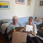Stariji muškarac i žena sjede na kauču u svom domu i pokazuju paket kućnih potrepština koji su dobili u sklopu projekta „Zaželi SUNCE”.
